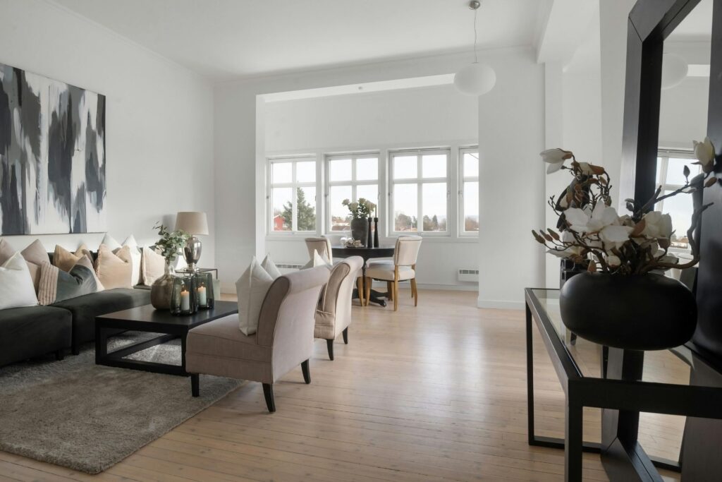 A modern living room with neutral furniture, abstract wall art, a coffee table with decorative items, engineered hardwood installation, and a dining area by large windows.