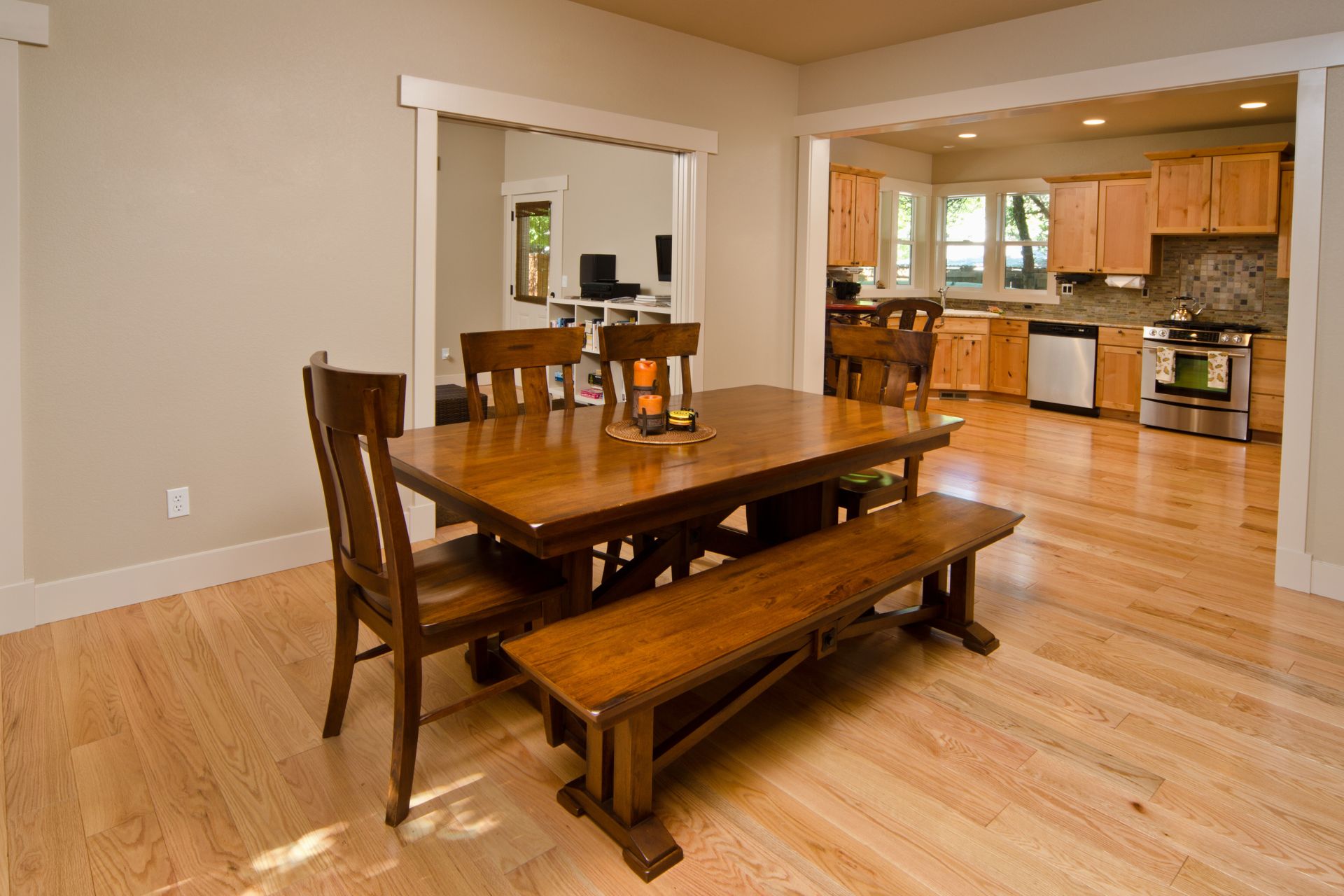 A wooden dining table with chairs and a bench sits on an engineered hardwood installation, adjacent to an open kitchen with light wood cabinets and stainless steel appliances.