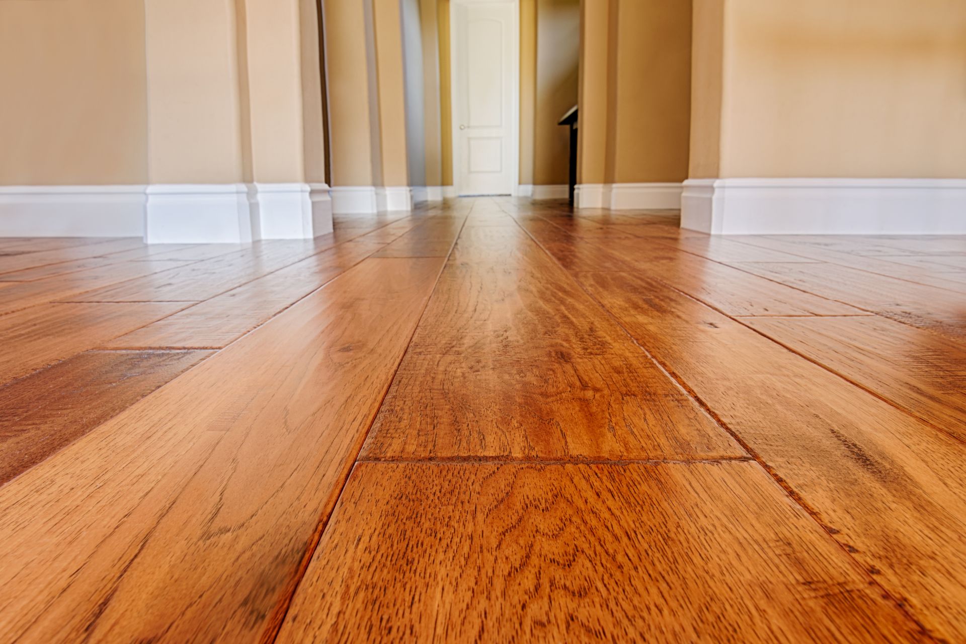 Close-up view of polished floorboards after engineered hardwood installation in a hallway, with cream-colored walls, white baseboards, and a closed door in the background.