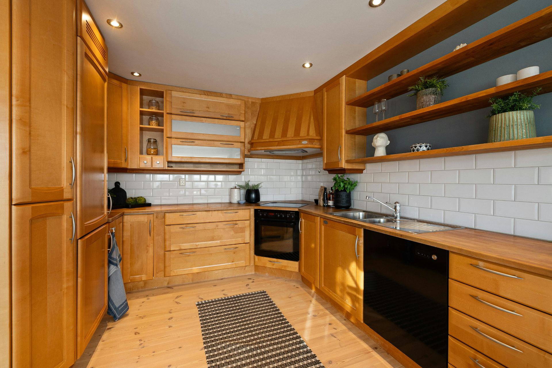 A modern kitchen with engineered hardwood installation, wooden cabinets, black appliances, white subway tile backsplash, open shelves, plants, and a striped rug on a wood floor.