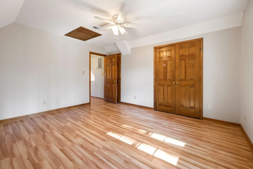 Empty room with engineered hardwood installation, neutral walls, a ceiling fan, wooden doors, baseboards, and sunlight streaming in from an unseen window.