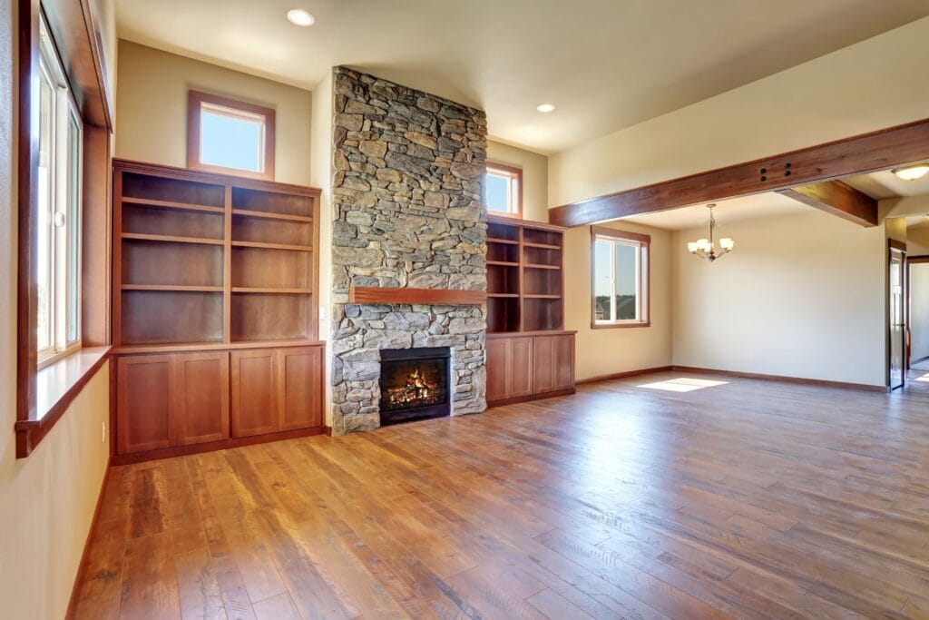 Empty living room with engineered hardwood installation, built-in wooden bookshelves, and a stone fireplace centered between two tall windows.