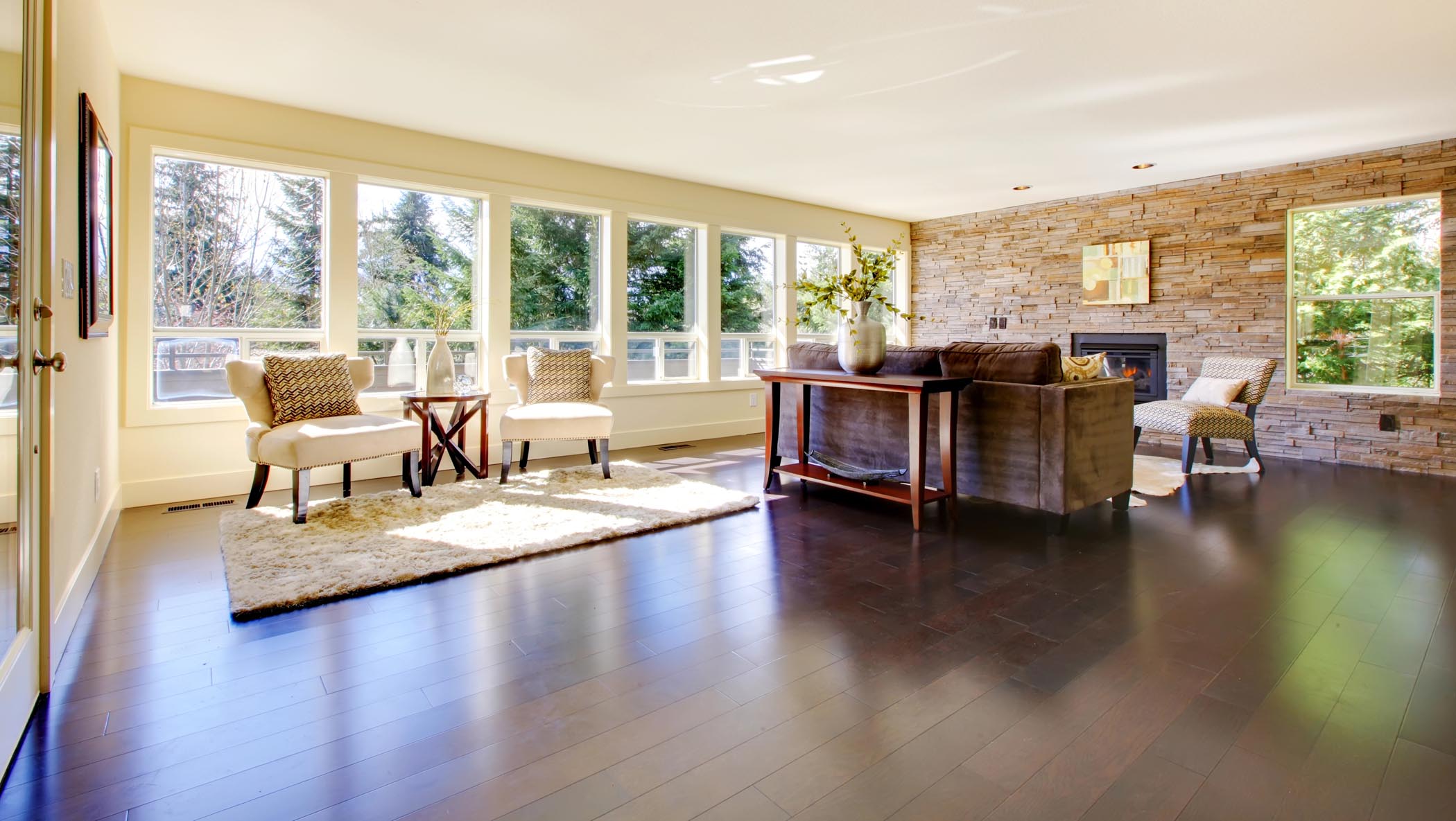 Spacious living room with dark wood floors, large windows, stone fireplace, neutral furniture, and a decorative rug. Natural light fills the space.