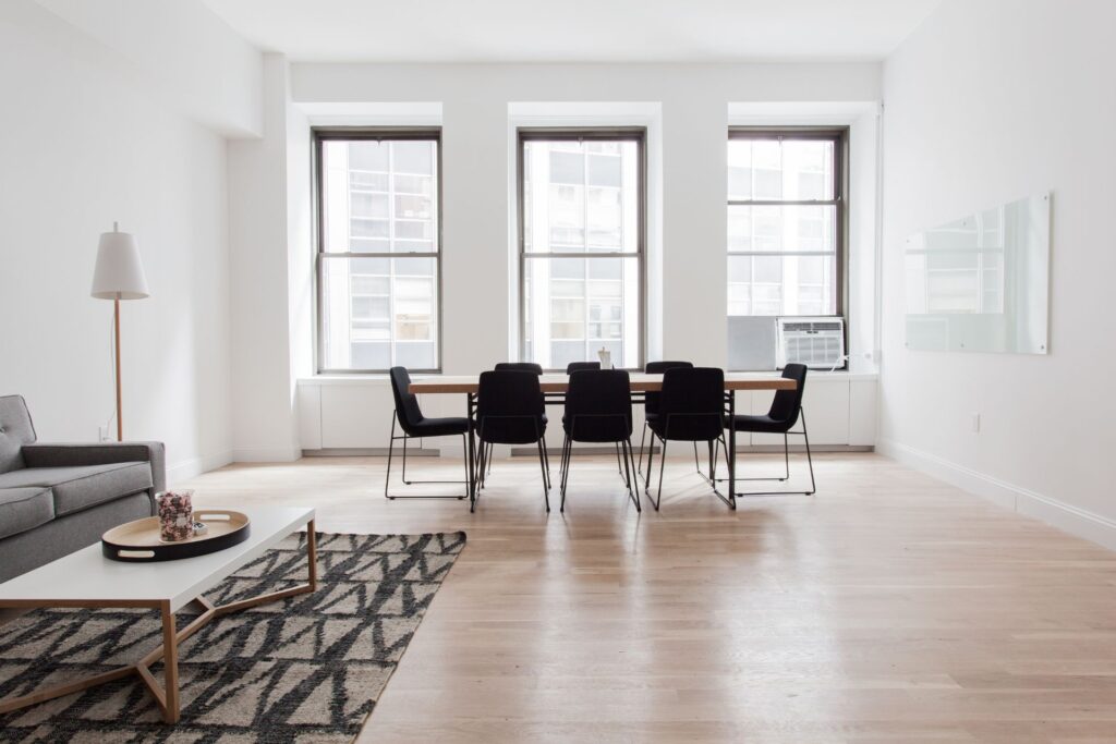A modern, minimalist room with a long table and six chairs near three large windows features expert flooring installation in Georgia, complemented by a gray sofa, a white floor lamp, and a coffee table on a patterned rug.