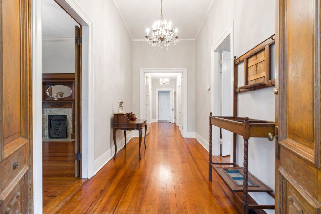 A hallway with polished wooden floors—showcasing expert flooring installation in Georgia—light-colored walls, chandeliers, a small table, and a wooden cart, leading to multiple white doors at the far end.