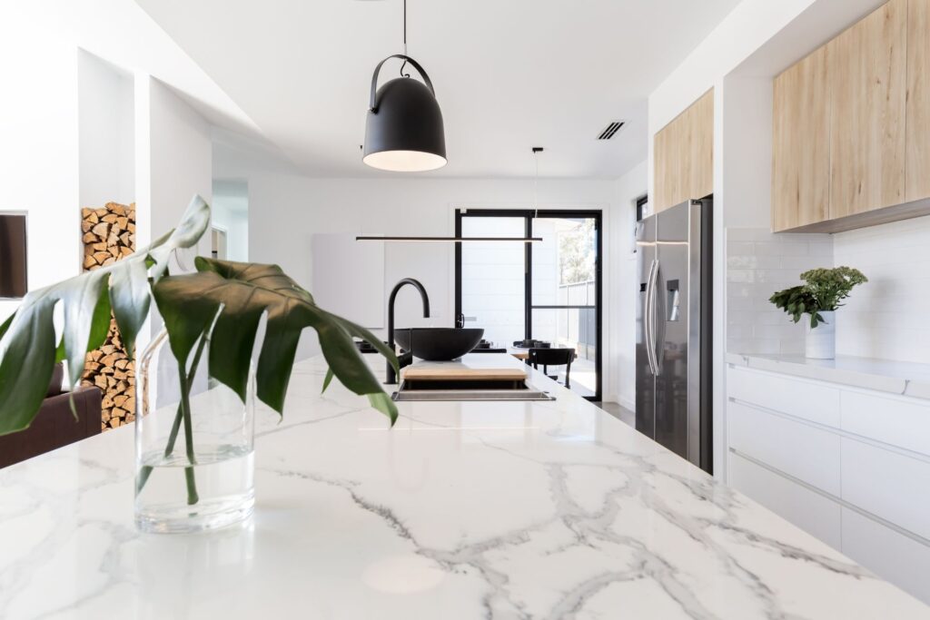 Modern kitchen with a white marble island, black pendant lights, stainless steel appliances, light wood cabinets, and sleek laminate flooring; a green plant in a glass vase sits on the counter.