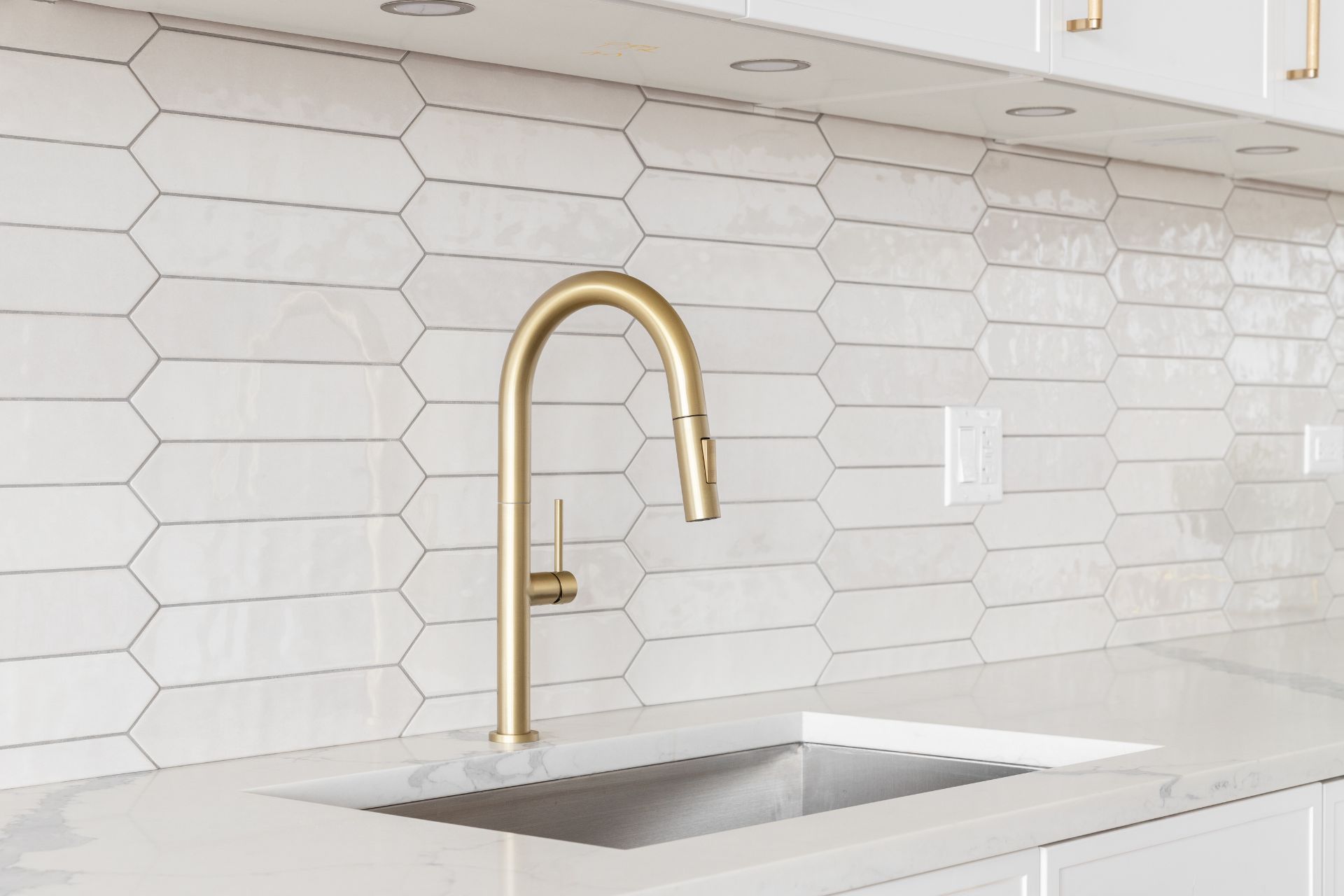 A modern kitchen sink with a gold faucet set in a white marble countertop, featuring a white hexagonal tile backsplash and complemented by warm laminate flooring.