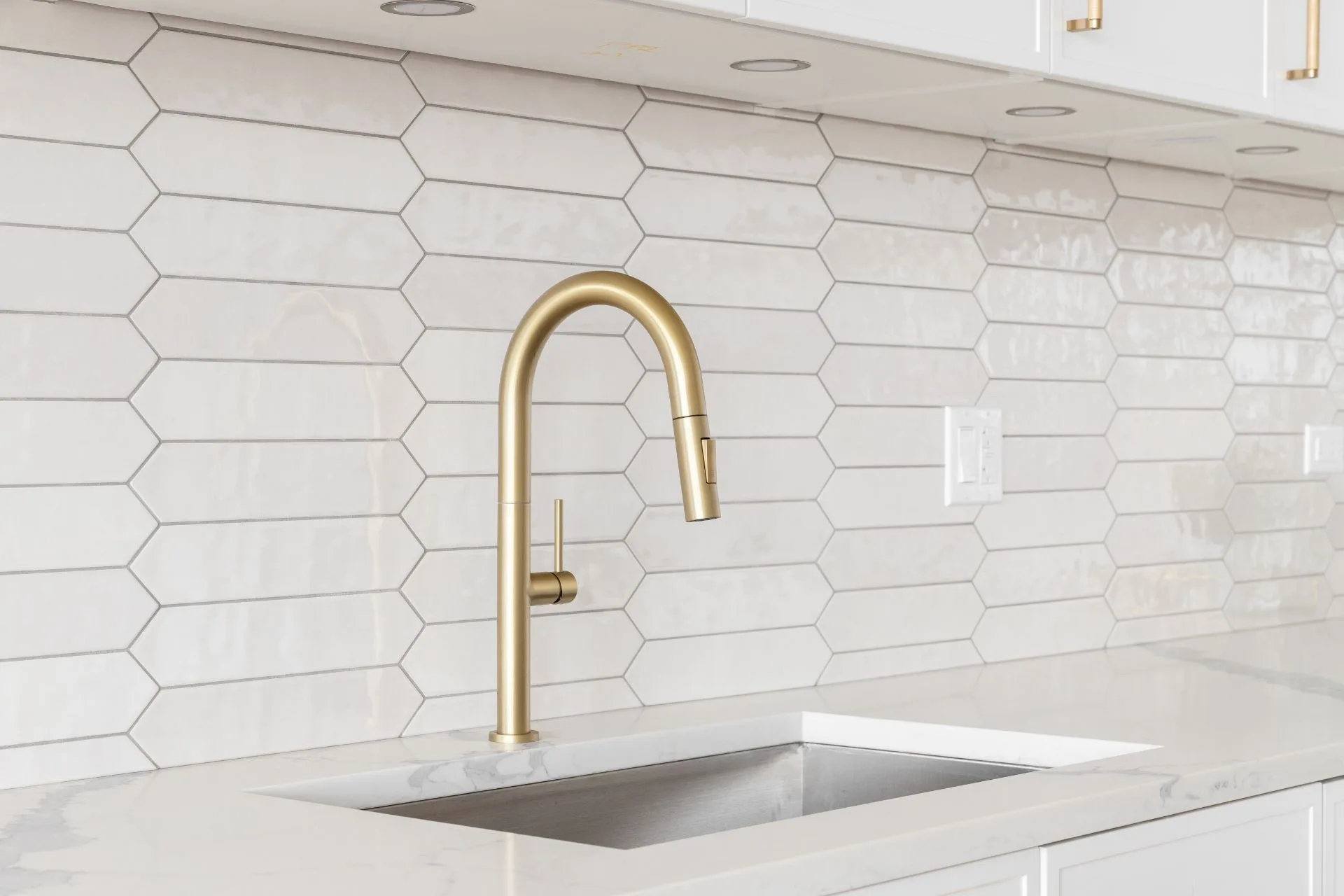 A modern kitchen sink with a gold faucet set in a white marble countertop, featuring a white hexagonal tile backsplash and complemented by warm laminate flooring.