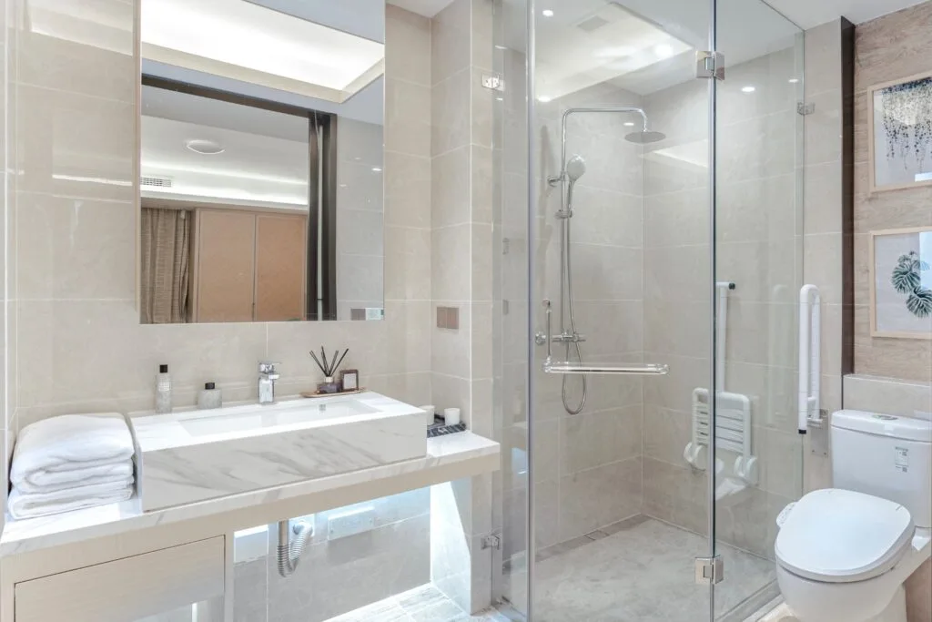 Modern bathroom with laminate flooring, a glass-enclosed shower featuring expert tile installation in Georgia, a wall-mounted toilet, large sink with marble countertop, and neatly folded towels.