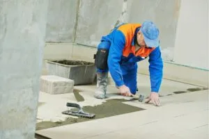 A construction worker in blue uniform and cap, known as the best tile contractor in Marietta, installs ceramic floor tiles in a room using a rubber mallet and adhesive.