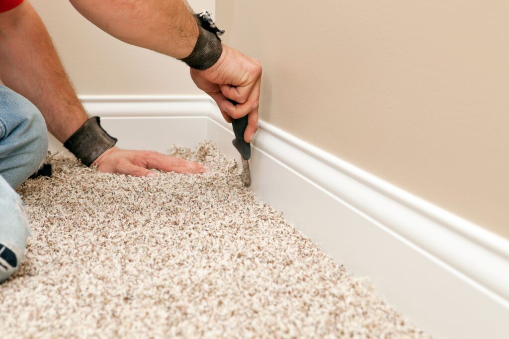 A person uses a tool to tuck carpet edges under white baseboard molding along a beige wall, demonstrating precise carpet installation techniques.