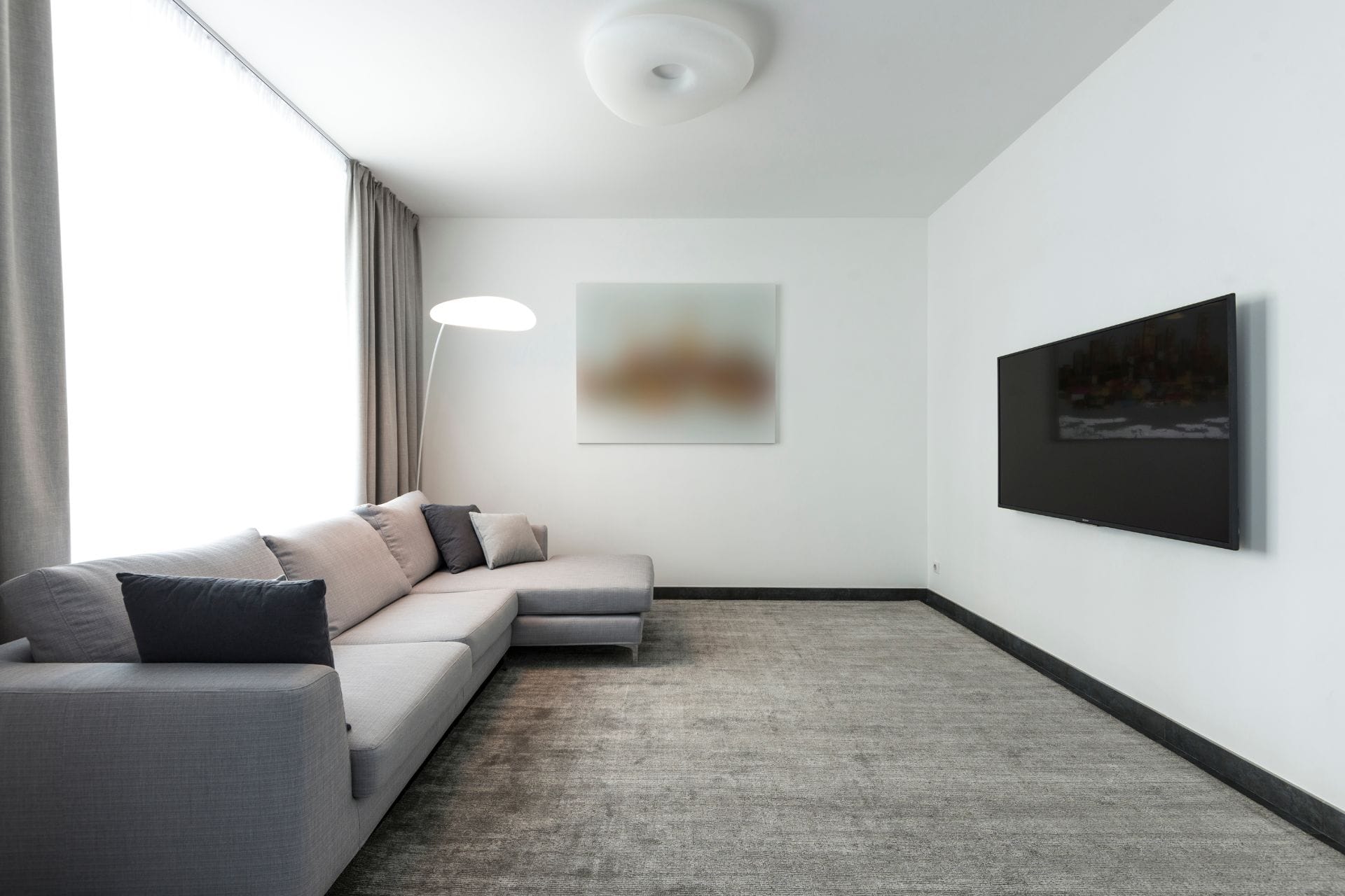 Minimalist living room with a gray sectional sofa, floor lamp, large wall-mounted TV, blurred wall art, and expertly done carpet installation featuring a sleek gray carpet.