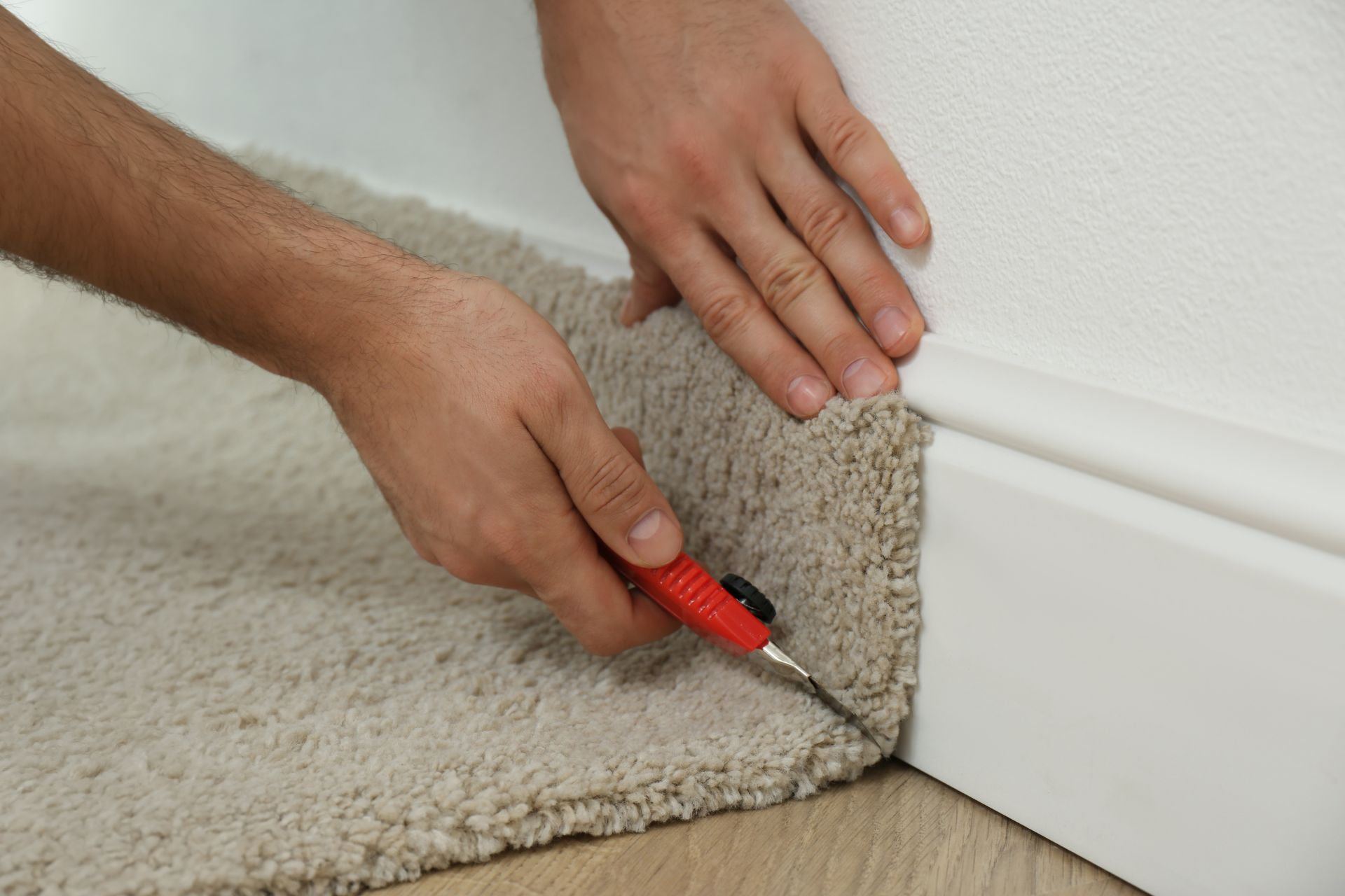 A person cuts beige carpet along the edge of a white wall and baseboard using a red utility knife during carpet installation.
