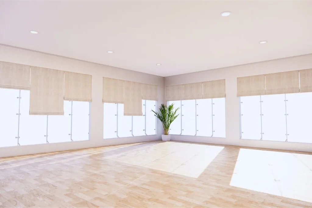 A spacious, empty room with light wood flooring, large windows with beige blinds, and a potted plant in the corner.