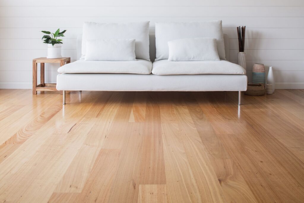 A minimalist living room with a white sofa, light wood flooring, a small side table with a plant, and decorative items against a white shiplap wall.