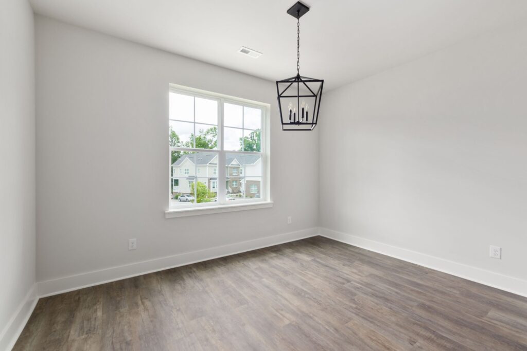 Empty room with white walls, wood flooring, a large window showing neighboring houses, and a black geometric ceiling light fixture.
