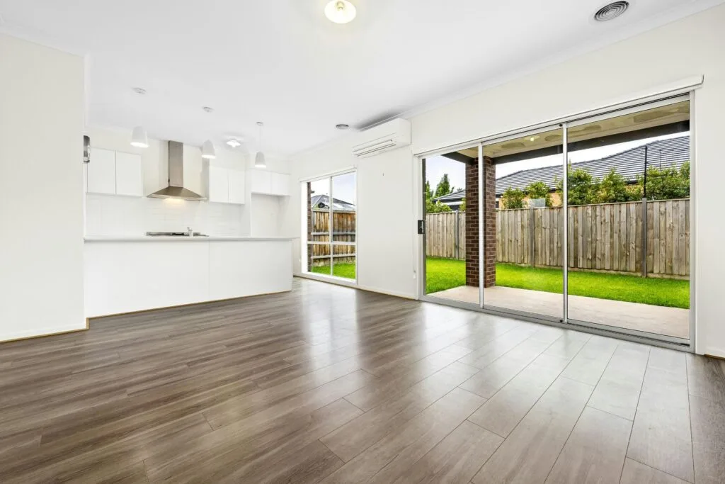 Abel Pro Flooring Gallery: A modern, empty open-plan living area with wood flooring, a white kitchen, large windows, and sliding glass doors leading to a fenced backyard with green grass.