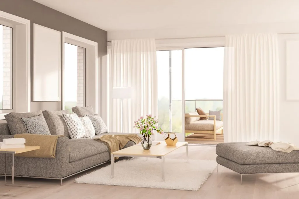 Modern living room with gray furniture, a coffee table with a vase of flowers, large windows with white curtains, and an outdoor seating area visible through sliding doors.
