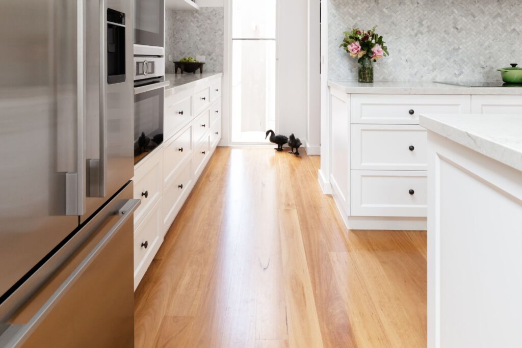 Modern kitchen with white cabinets, stainless steel appliances, wooden floor, and a black cat near a glass door. A vase of flowers is on the counter.