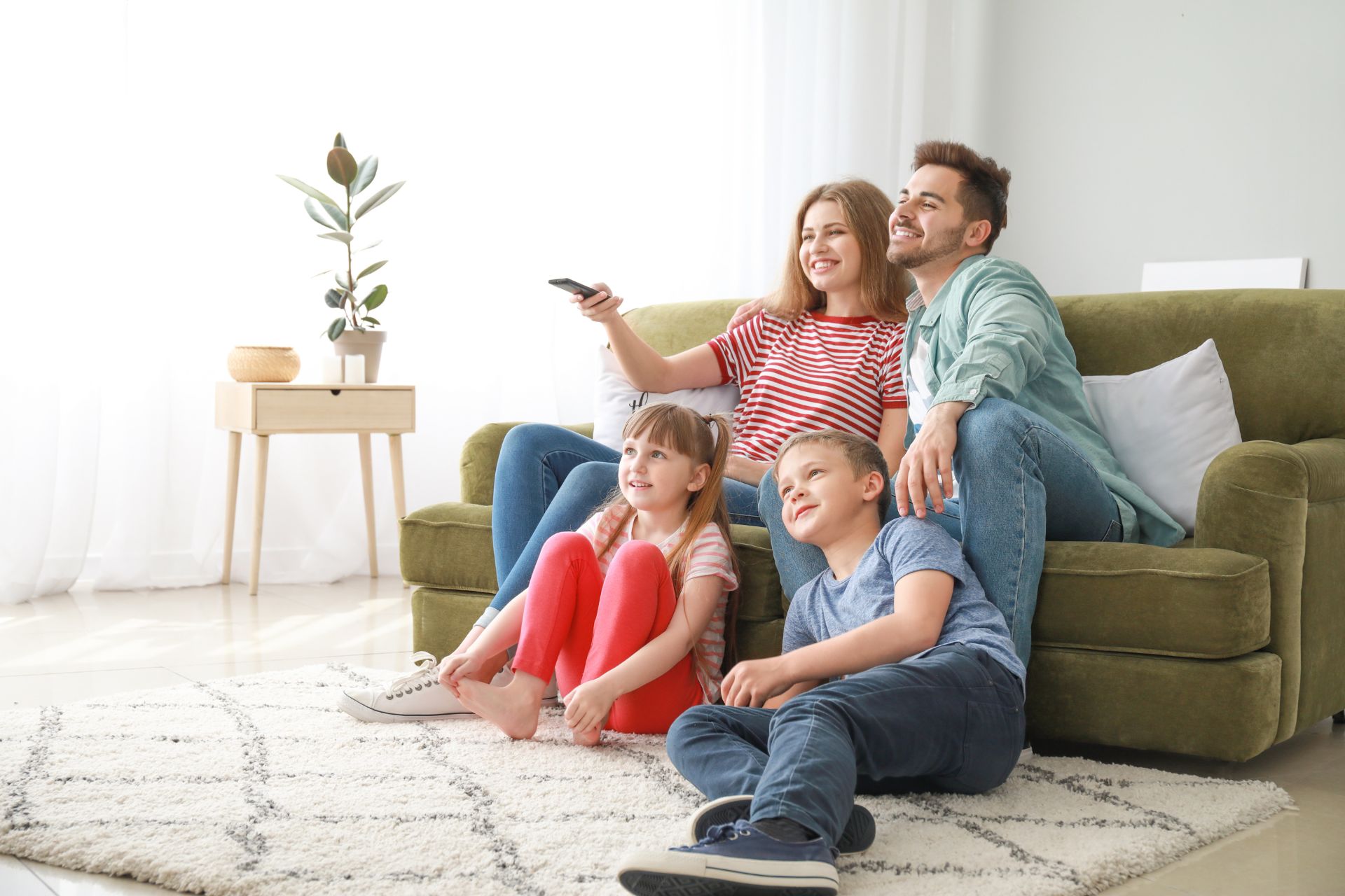 A smiling family of four sits together in a living room, watching TV. The parents sit on a green sofa while two children sit on the carpet in front of them.