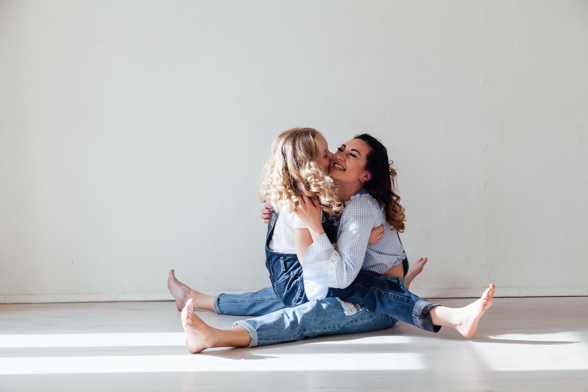 Two people sitting on a light floor, facing each other with legs crossed, hugging and smiling in a bright, minimalist room.