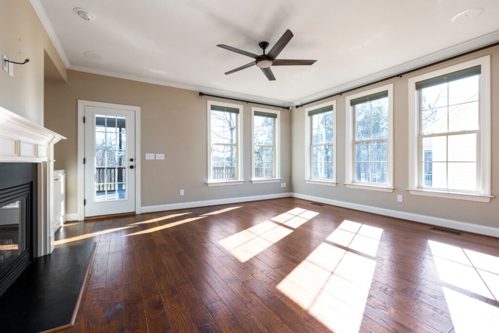 Bright empty living room with hardwood floors, large windows, a ceiling fan, a fireplace, and a door leading to an outdoor deck.
