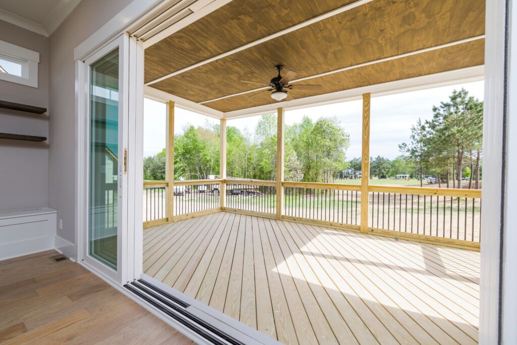 Open sliding glass doors leading to a covered wooden deck with a ceiling fan, surrounded by railings and overlooking a grassy area with trees in the background.