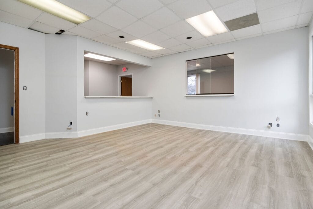 Empty office space with laminate flooring in a light wood finish, white walls, a ceiling with fluorescent lights, a large window, and a service counter with an open doorway in the background.