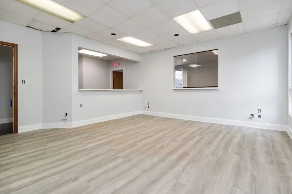 Empty office space with laminate flooring in a light wood finish, white walls, a ceiling with fluorescent lights, a large window, and a service counter with an open doorway in the background.