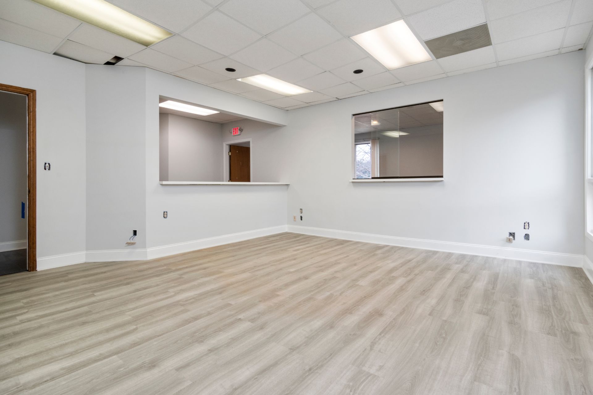 Empty office space with laminate flooring in a light wood finish, white walls, a ceiling with fluorescent lights, a large window, and a service counter with an open doorway in the background.