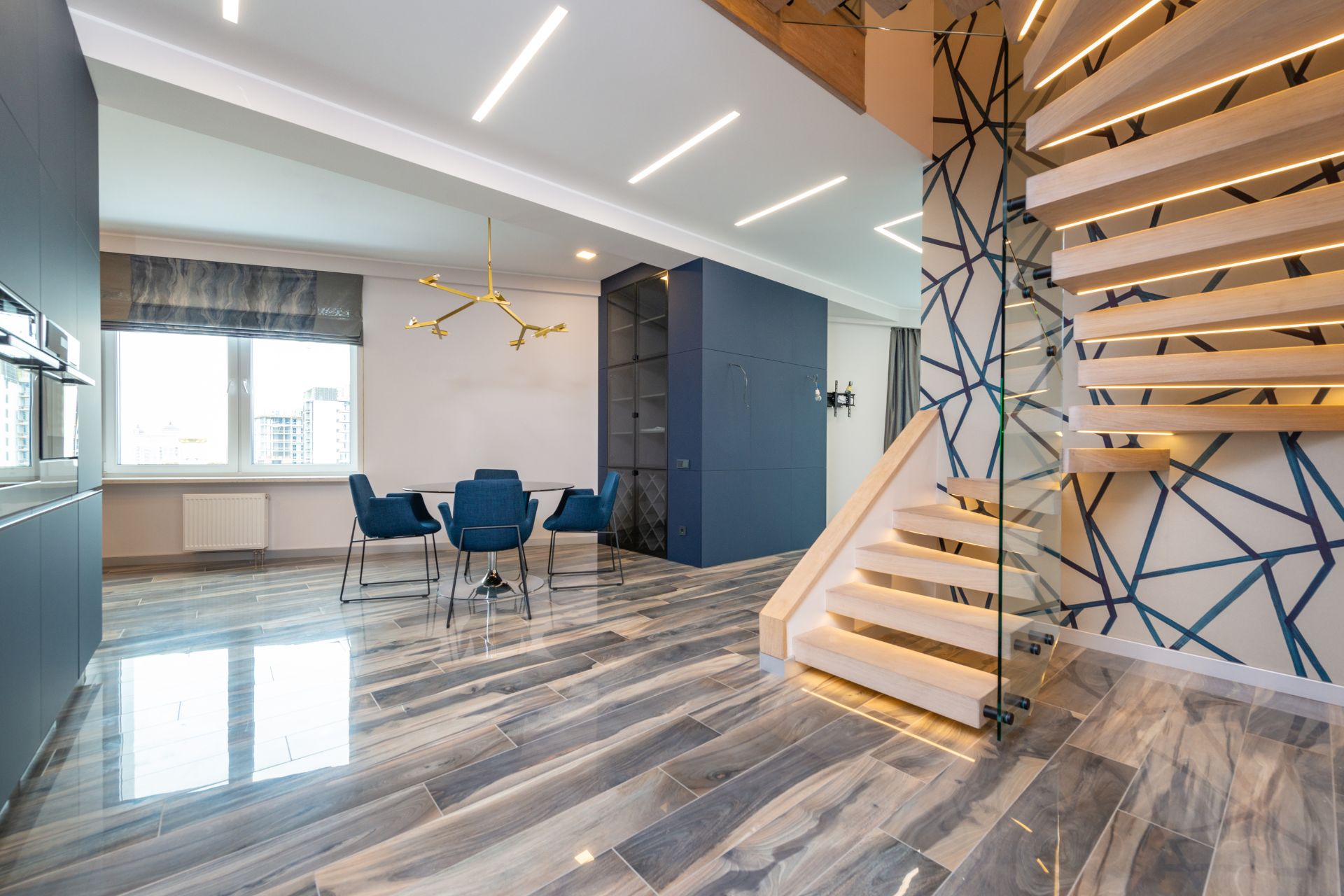 Modern open-plan interior with a wooden floating staircase, geometric wall design, dining area with blue chairs, large window, and sleek marble-patterned laminate flooring, showcasing expert laminate flooring installation in Georgia.