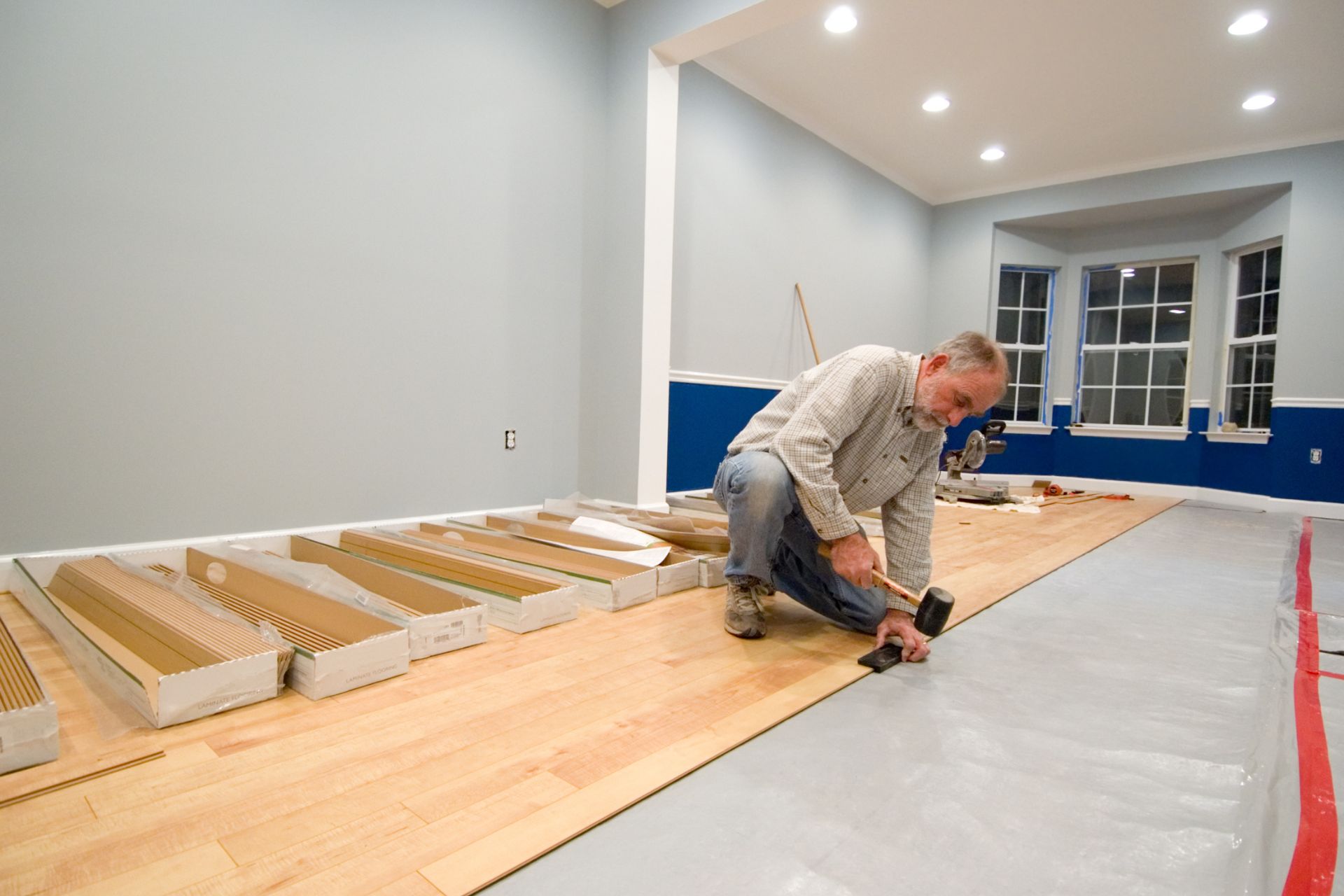 A man uses a mallet to install laminate flooring in a brightly lit room, with boxes of planks and tools on the unfinished section, showcasing details about Abel Pro Flooring.