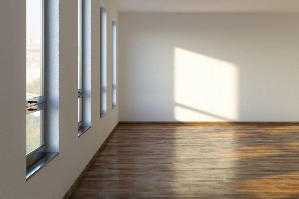 Empty room with light-colored walls, laminate flooring, and four tall windows on the left letting in sunlight that casts a square pattern on the wall and floor.