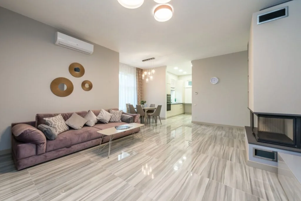 Modern living room with a mauve sofa, decorative wall mirrors, dining area, and open view into a bright kitchen. Laminate flooring with a marble-like finish and a built-in fireplace are visible, showcasing excellent LVP Installation in Georgia.