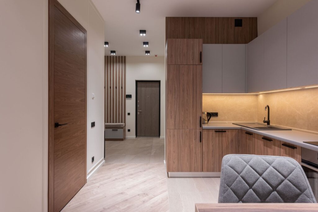 Modern minimalist kitchen and hallway with wood accents, light walls, built-in cabinets, a closed door, beige backsplash, laminate flooring, and overhead lighting.