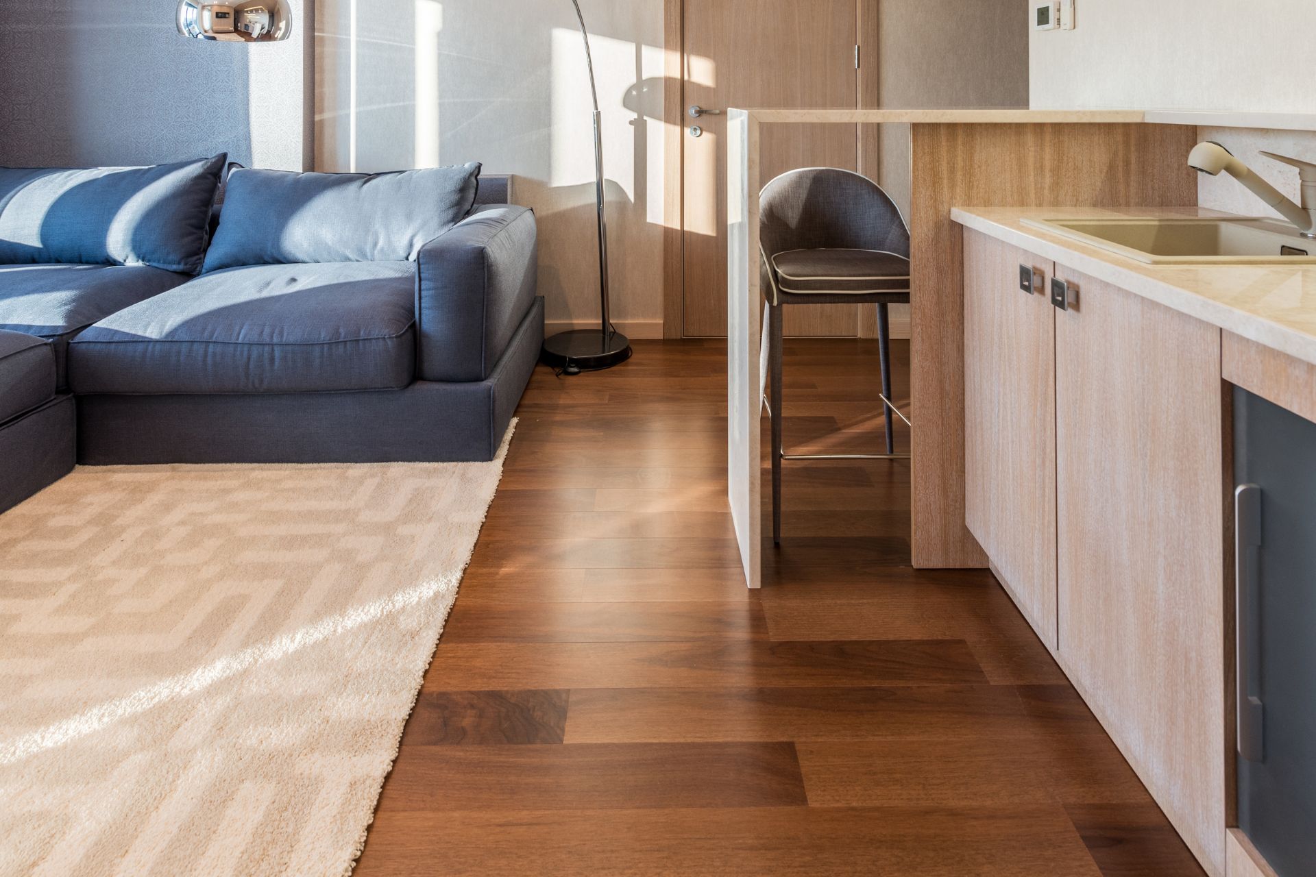 Modern living space with a blue sectional sofa, beige rug, stylish laminate flooring, kitchenette with light wood cabinets, bar stool, and standing lamp.
