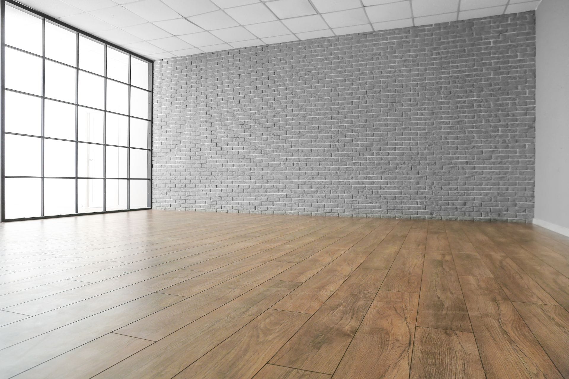 Empty room with a large grid window, gray brick wall, ceiling tiles, and light brown laminate flooring.