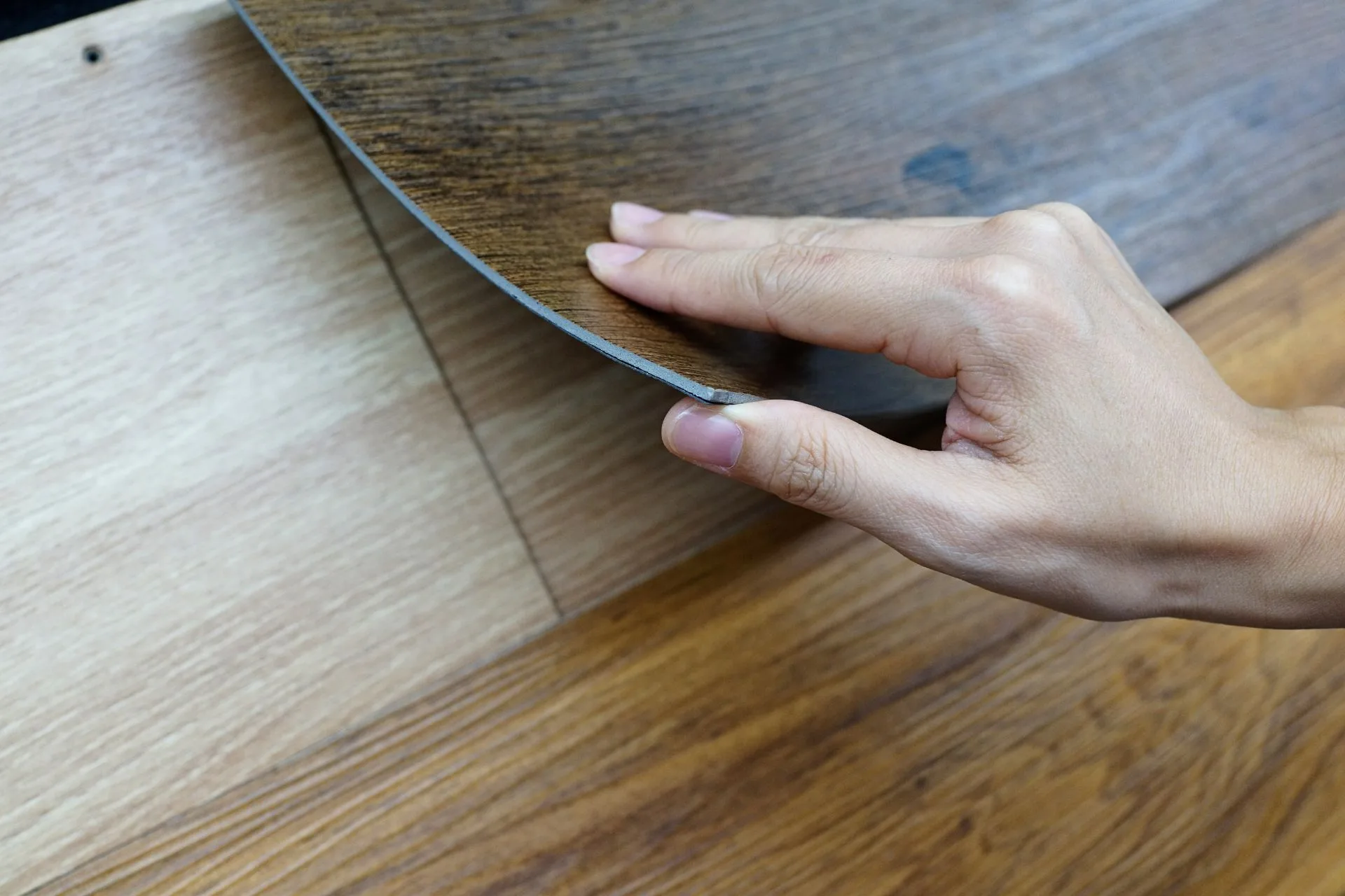 A hand demonstrates VCT flooring installation in Georgia, placing a peel-and-stick vinyl flooring plank with a dark wood-look finish over a lighter wood-look surface.