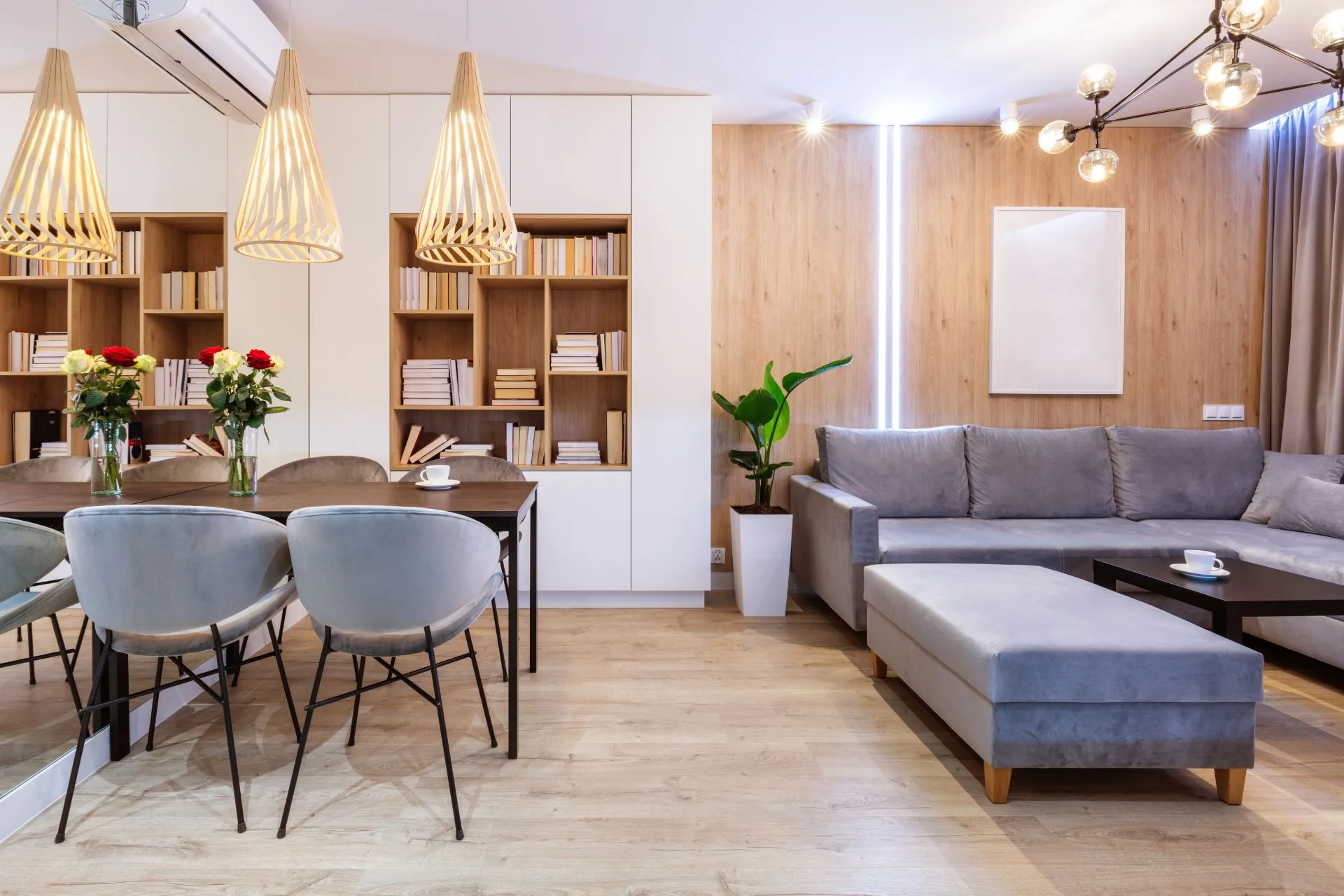 Modern living and dining area with grey sectional sofa, wooden dining table, four velvet chairs, pendant lights, bookshelves, and potted plants on luxury vinyl plank flooring.