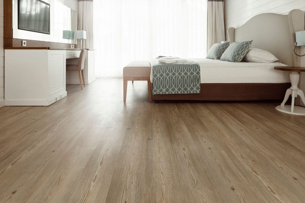 A modern bedroom with a large bed, luxury vinyl plank flooring installation, desk, wall-mounted TV, and floor-to-ceiling windows with sheer curtains letting in natural light.