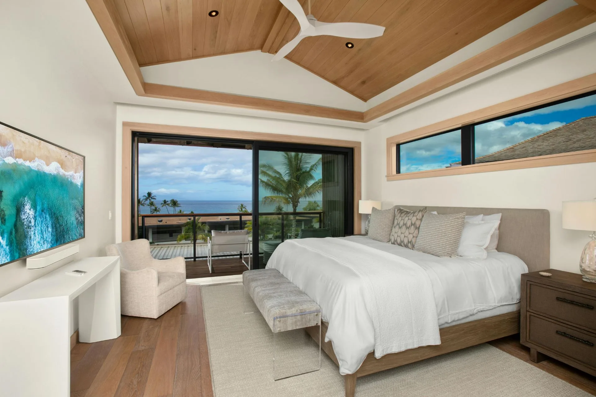 Modern bedroom with a large bed, wooden ceiling, white walls, and sliding glass doors opening to an ocean-view balcony. Luxury vinyl plank flooring adds elegance, while a TV and armchair are situated on the left side of the room.