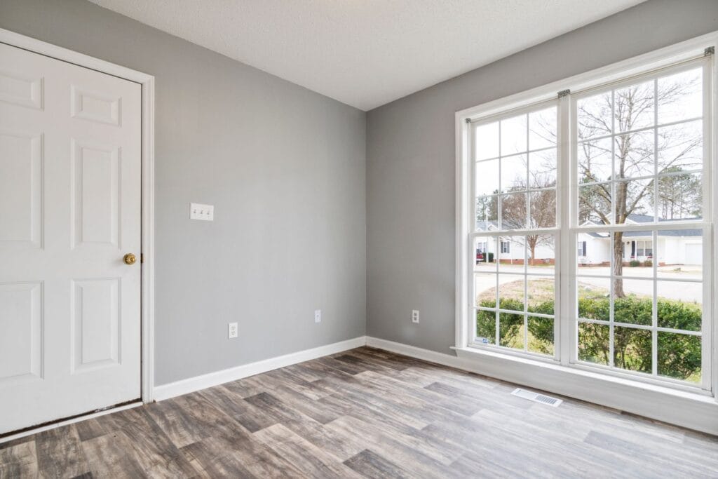 A small room with gray walls, luxury vinyl plank flooring, a white door, and a large window showing a view of the street and bushes outside.