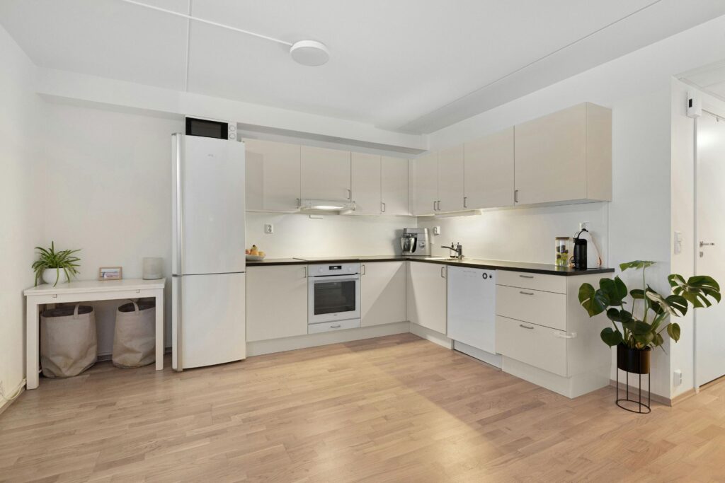 Modern L-shaped kitchen with white cabinets, built-in appliances, and expert Engineered Hardwood Flooring Installation in Georgia for the light wood flooring. A refrigerator, houseplants, and storage bins complete the minimal decor.