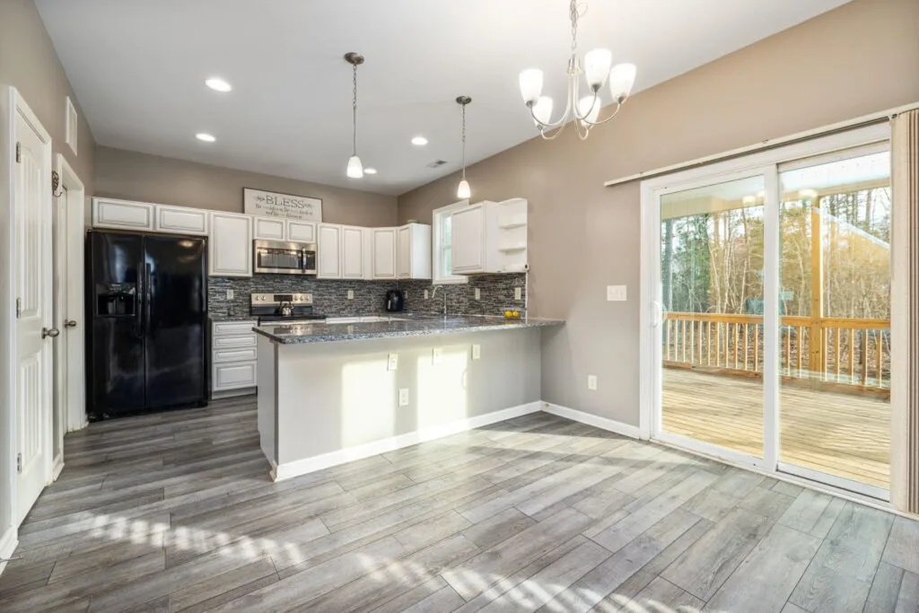 Modern kitchen with white cabinets, stainless steel appliances, gray luxury vinyl plank flooring, and sliding glass doors leading to a wooden deck with a view of trees outside.