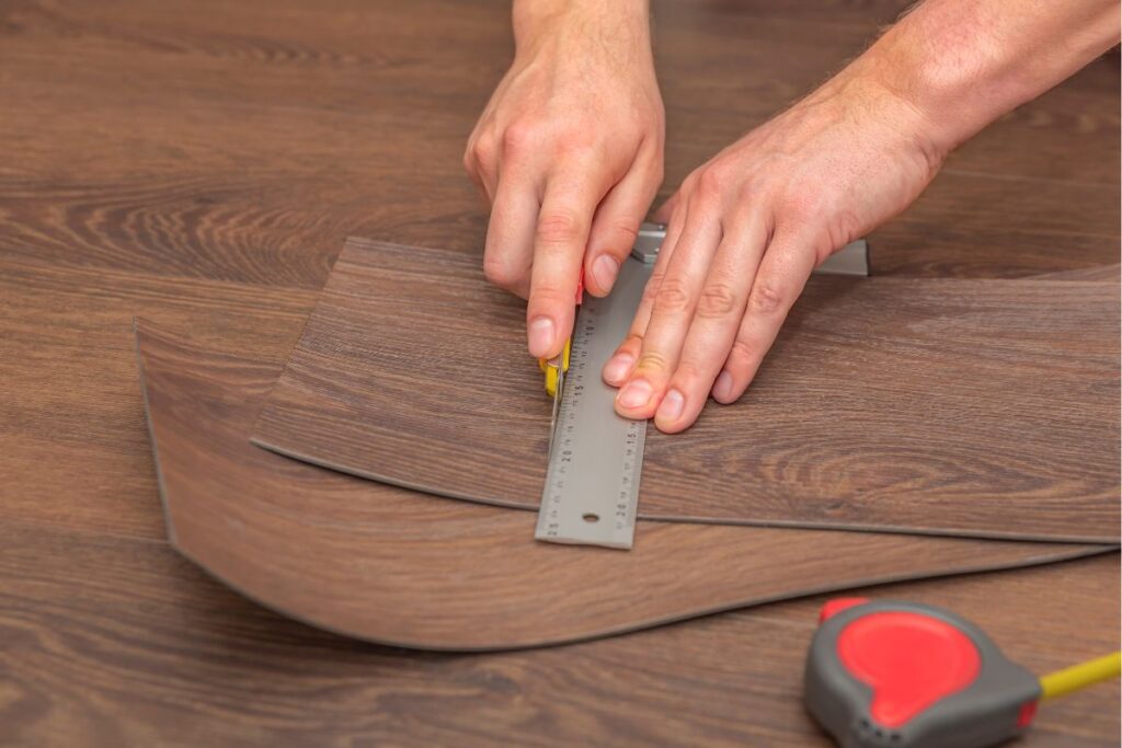 A person uses a metal ruler and utility knife to cut wood-patterned vinyl flooring—preparing materials while considering LVP installation cost in Georgia; a tape measure is on the table.