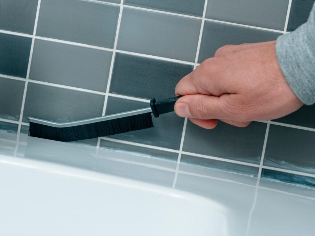 Hand using a grout brush to clean tile grout lines in a bathroom setting, emphasizing effective grout cleaning techniques for maintaining tile surfaces.