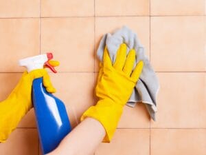 Person wearing yellow gloves cleaning beige tiled wall with a blue spray bottle in one hand and wiping with a gray cloth in the other, demonstrating effective grout cleaning tips for sparkling results.