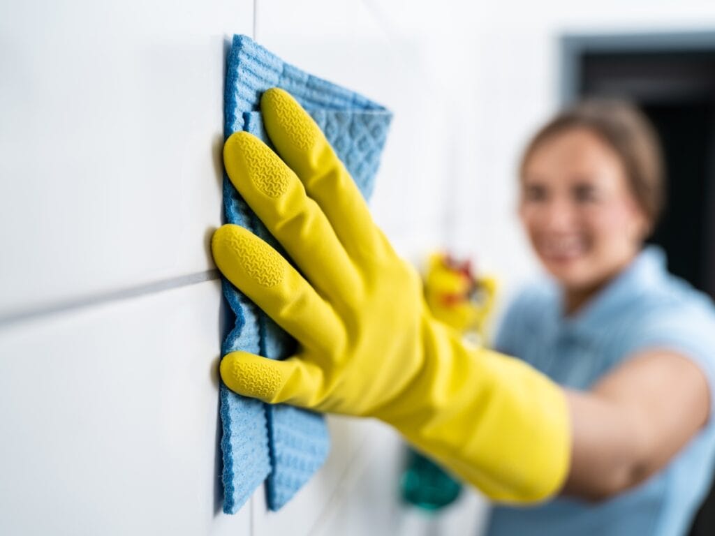 Person in yellow gloves cleaning grout with a blue cloth on white tiled wall, emphasizing effective tile maintenance and grout cleaning techniques.