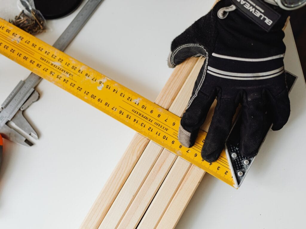 Hand in a black glove measuring wooden planks with a yellow ruler and square tool, illustrating essential tools for accurate flooring measurement.