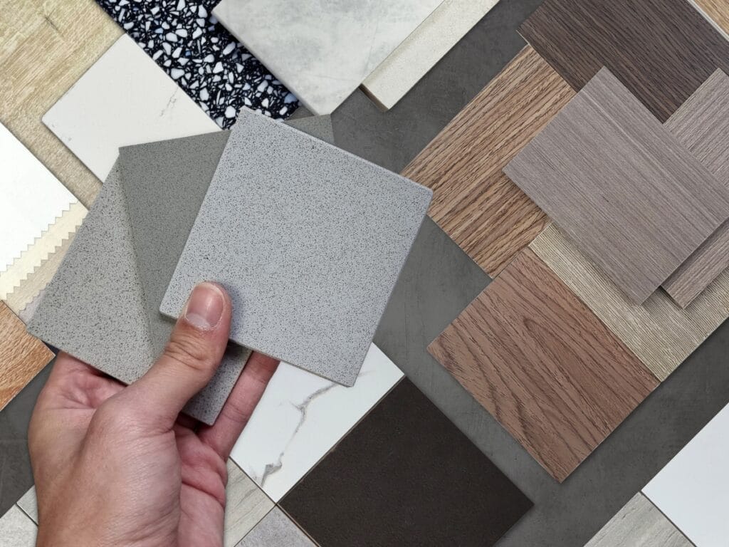 Hand holding various flooring samples, including gray textures, amidst a selection of different materials and colors, illustrating options for flooring measurement and installation.